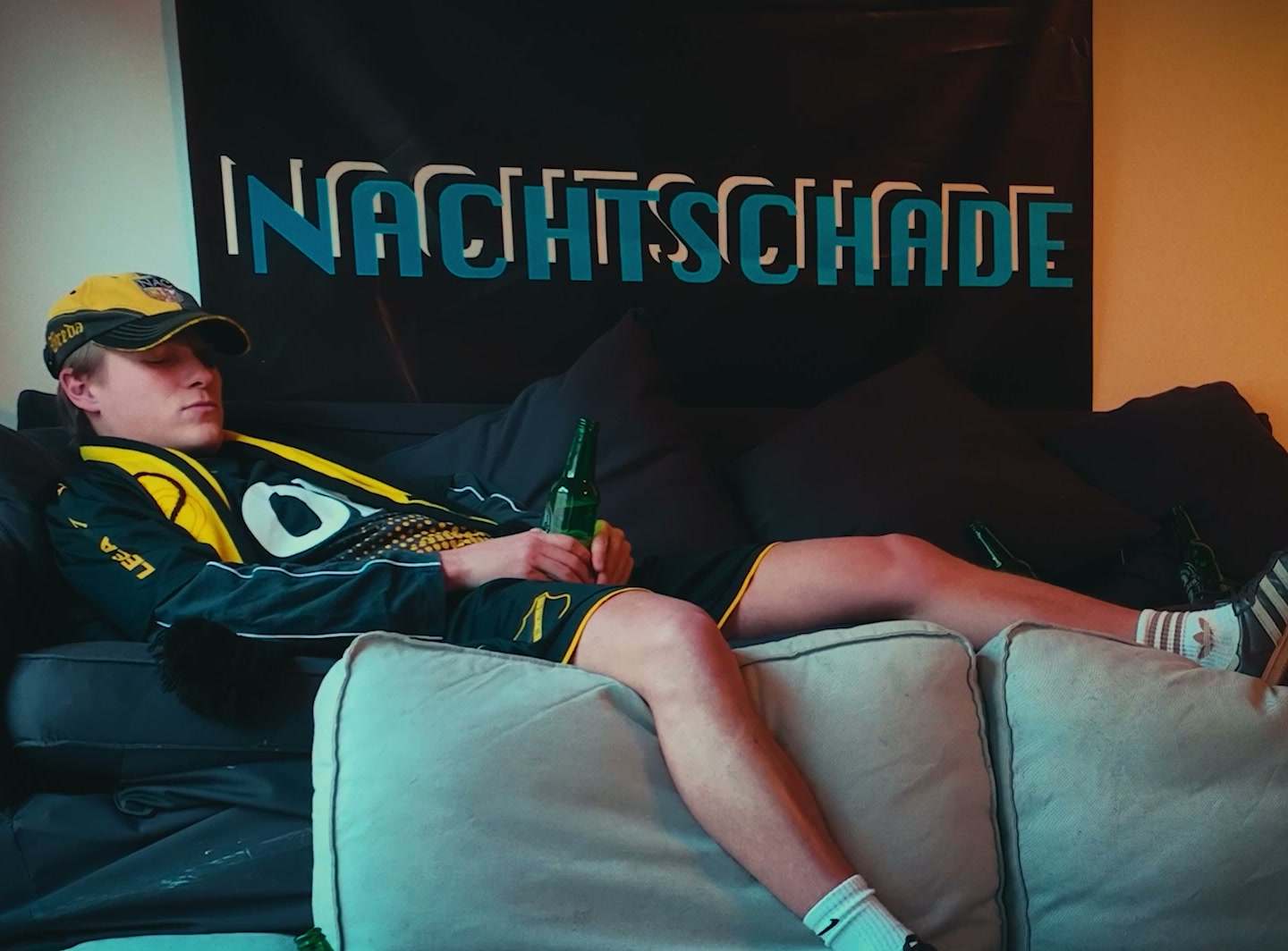 Een jongen in een geel-zwart voetbalshirt hangt op een bank onder een banner met de tekst Nachtschade.