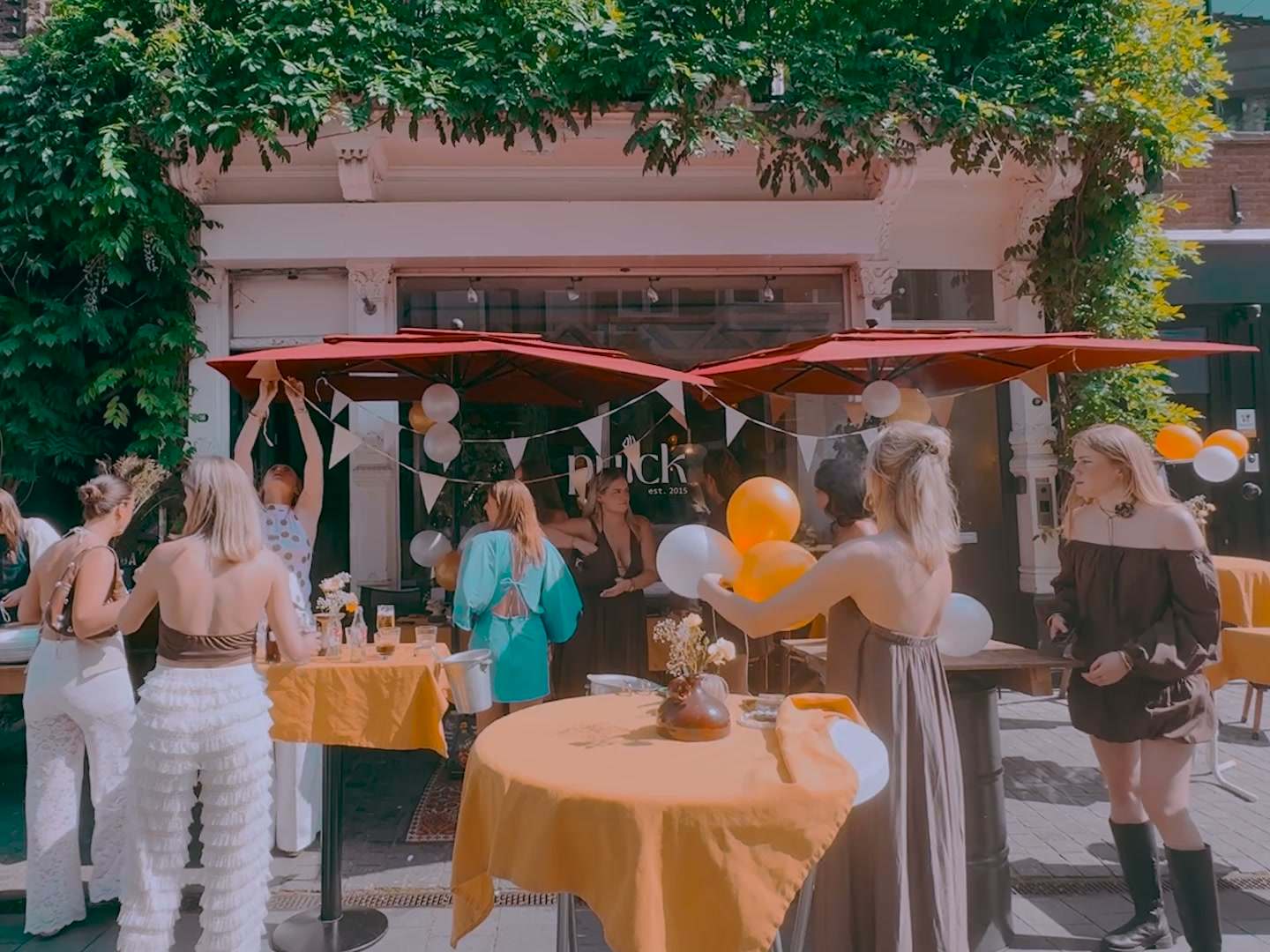 Een terras met rode parasols, vlaggetjes en oranje ballonnen, vol mensen in zomerkleding.