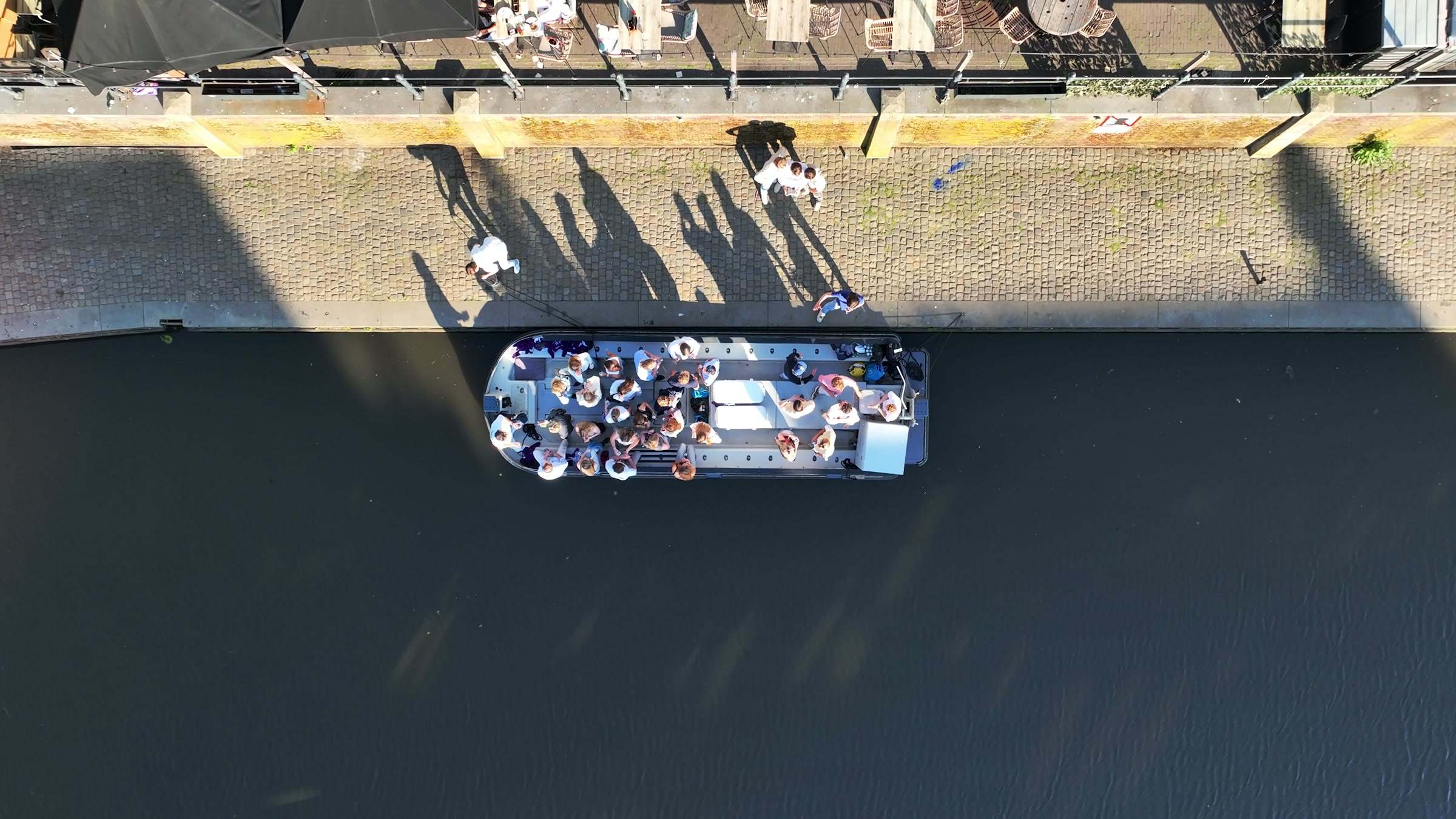 Luchtbeeld recht van boven van een sloep vol mensen op de gracht, langs een kade met bomen en terrassen.
