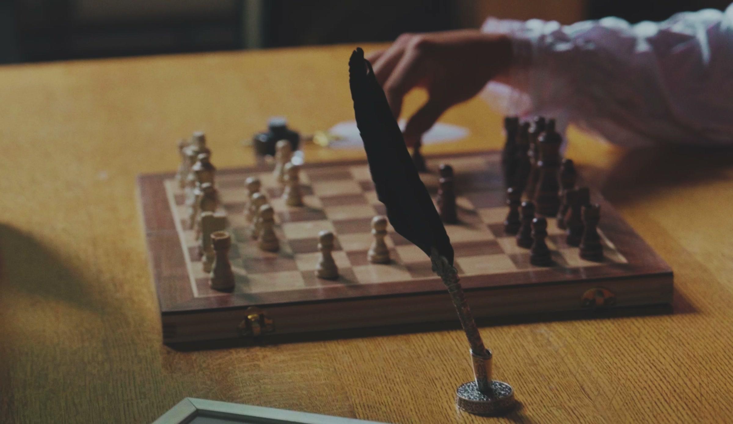 Een hand zet een schaakstuk op een houten bord, met een ganzenveer in een inktpot ervoor.